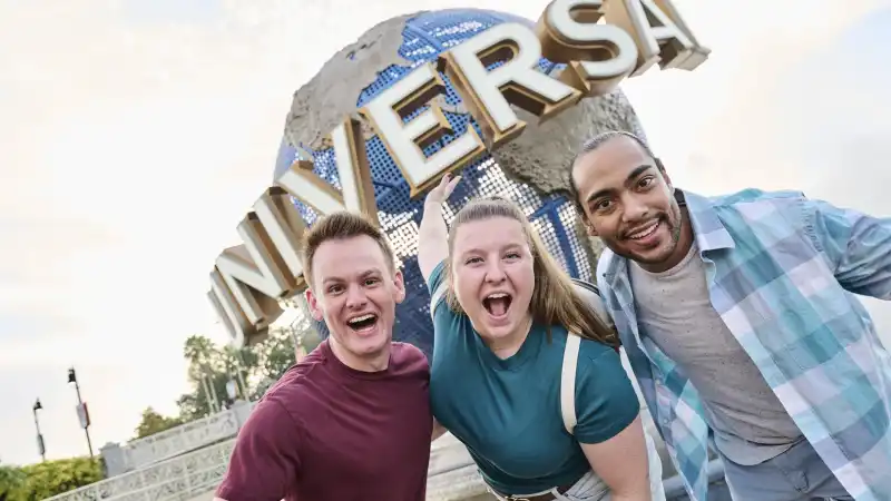 Three friends leaning in and cheering in front of the Universal globe at Universal Orlando Resort at sunset.