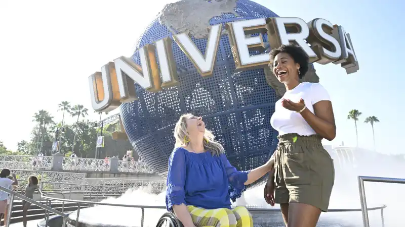Two women laughing together in front of the Universal globe fountain at Universal Orlando Resort, one of them seated in a wheelchair.