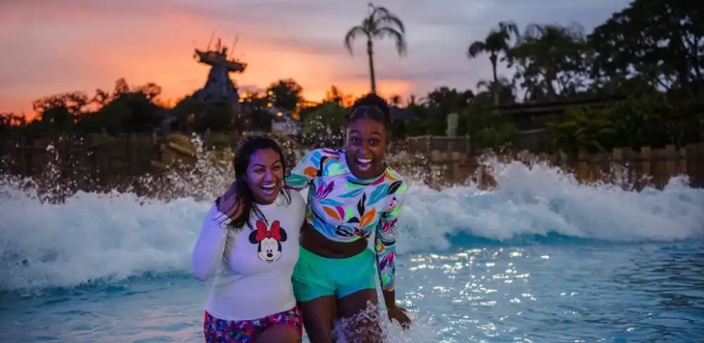 Two women smiling and enjoying themselves in a swimming pool at sunset with water splashing around them.