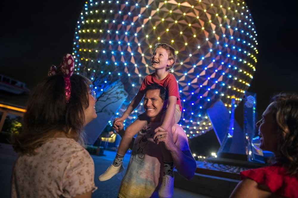A boy rides on his father's shoulders, both smiling in front of a brightly lit Spaceship Earth at EPCOT after dark.