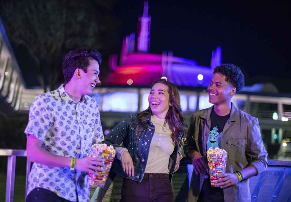 Three friends share popcorn beneath the illuminated peaks of Space Mountain at Magic Kingdom Park during an after-hours event.
