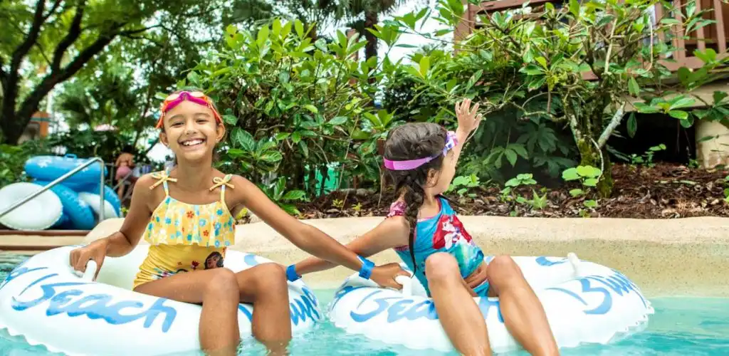 Two children smiling on inner tubes at the edge of a Disney water park pool surrounded by greenery