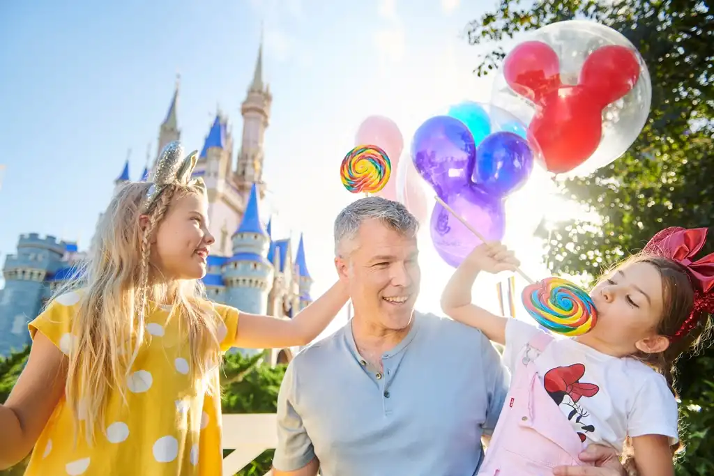A smiling man with two young girls at a theme park with a castle in the background, enjoying colorful balloons and lollipops on a sunny day.