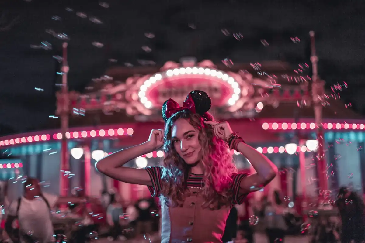 A guest in sparkling Minnie ears smiles in front of the glowing carrousel at Magic Kingdom Park at night.