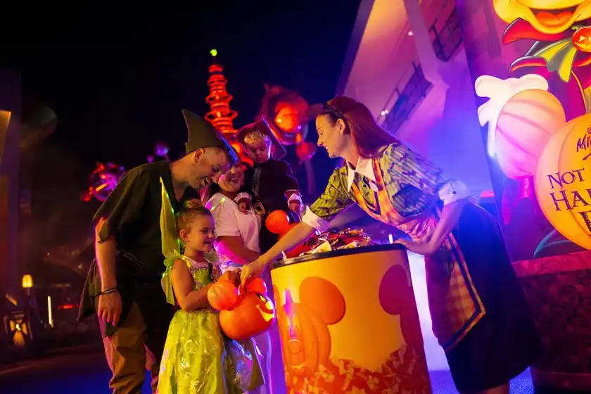 People in Halloween costumes celebrating with a hostess at a festive event with colorful pumpkin decorations.