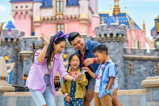 A family of four, smiling and laughing, looks at a mobile phone in front of Sleeping Beauty Castle at Disneyland. The parents and children are dressed in casual Disney-themed outfits, capturing a joyful moment together.