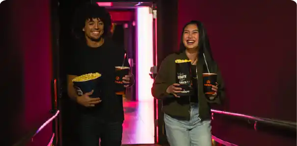 Two people stand in a dark hallway, each holding popcorn and drinks, ready for a movie experience.