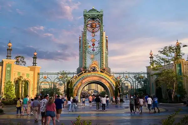 A majestic green-and-gold steampunk gateway topped by a clock tower and glowing “Epic Universe” sign, with guests milling across the plaza beneath a pastel dusk sky.