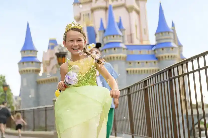 Young girl in a green princess dress and crown smiles while holding a bubble wand in front of a castle with blue spires at DisneyWorld