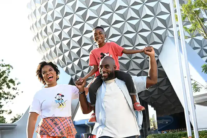 Happy family celebrating their visit to EPCOT, with a child on dad's shoulders and Spaceship Earth towering behind them
