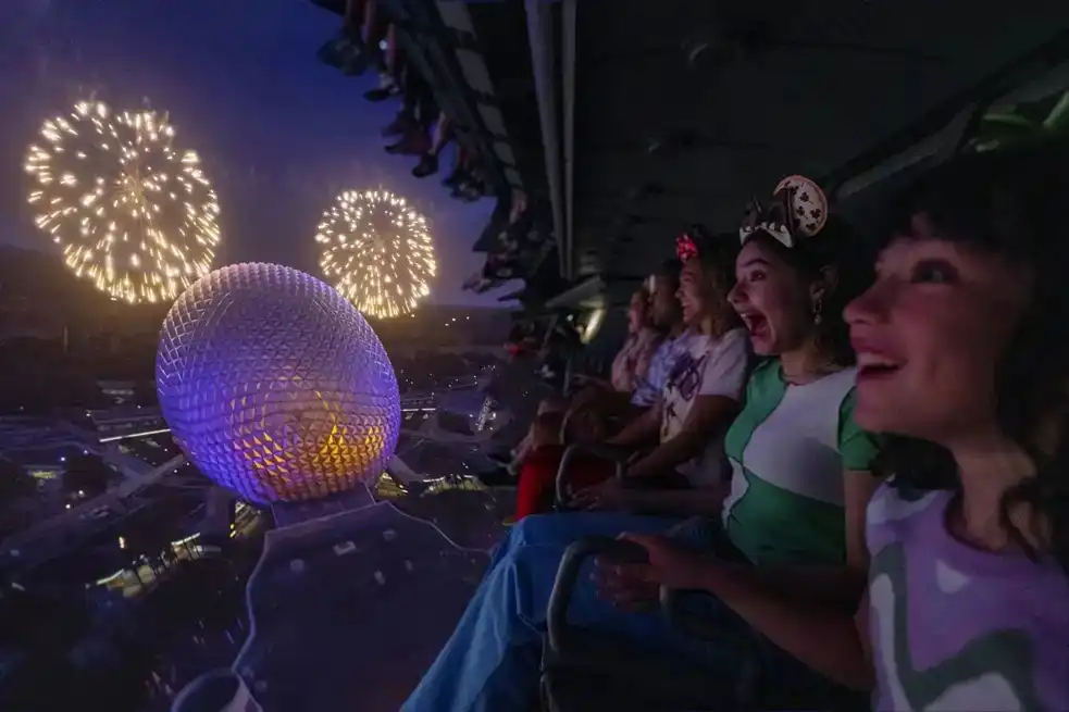 Guests experiencing the breathtaking flight of Soarin’ Around the World at EPCOT, soaring over Spaceship Earth with fireworks illuminating the sky. 