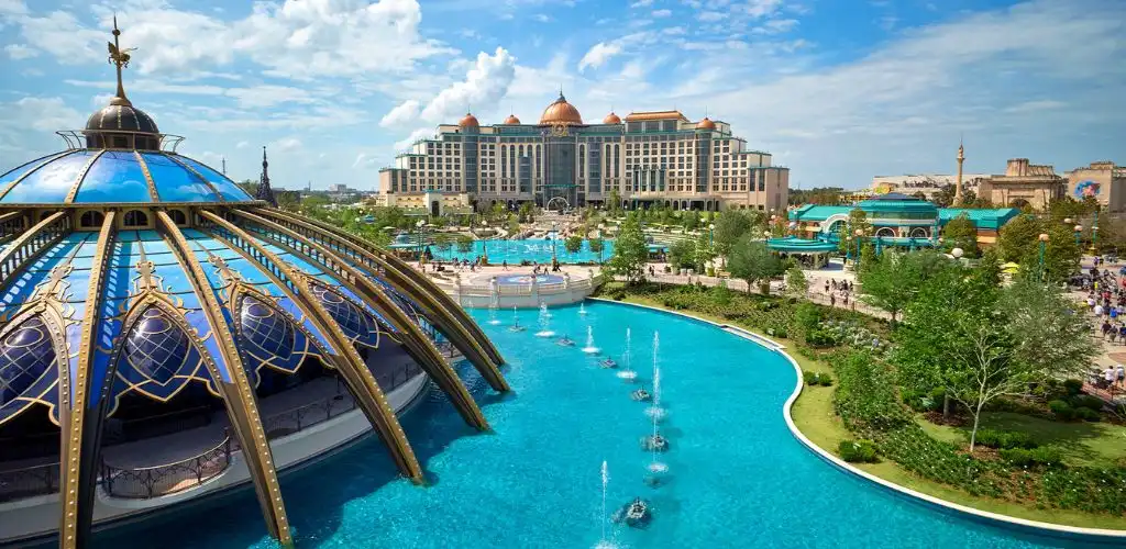 A view of a large resort with a domed building, water fountains, lush greenery, and a grand hotel in the background under a blue sky.