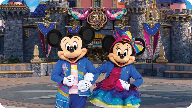 Mickey and Minnie Mouse dressed in colorful outfits standing happily in front of a castle decorated for a celebration.