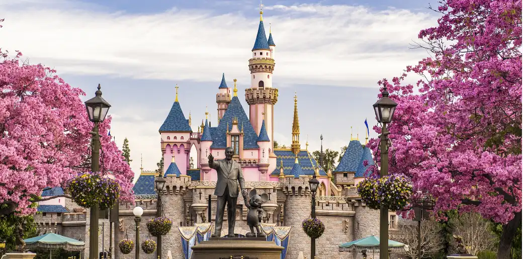 A pink and blue Disney castle with pink flowering trees and statues in front