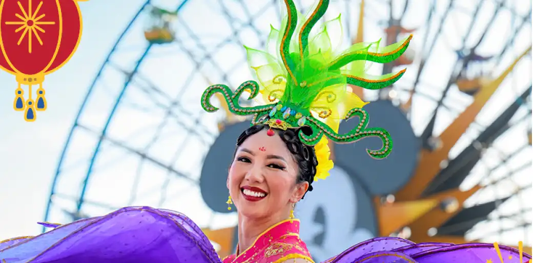 A smiling woman in colorful traditional costume with a green headpiece, standing in front of an amusement park ride and Ferris wheel during daytime.