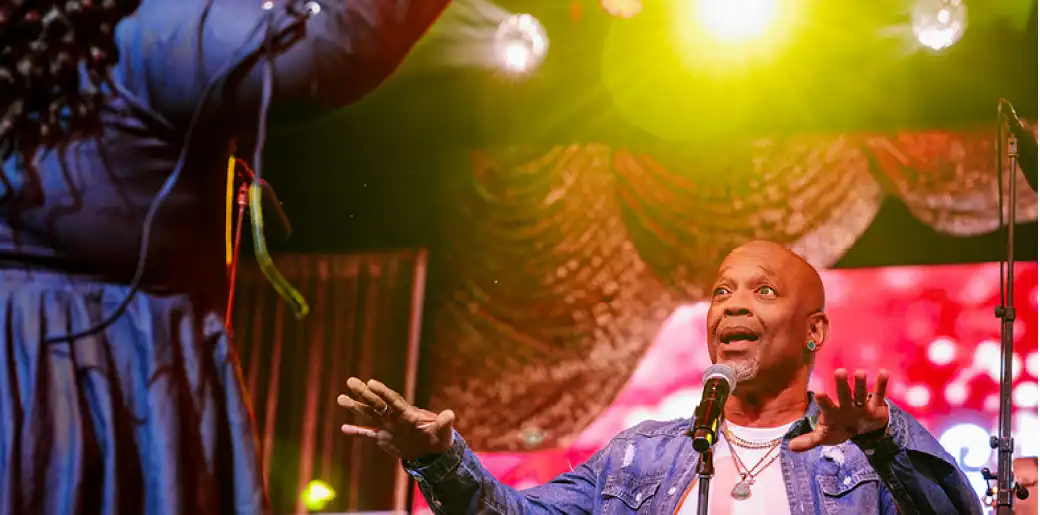 A performer singing into a microphone on stage with colorful lighting and curtains in the background.