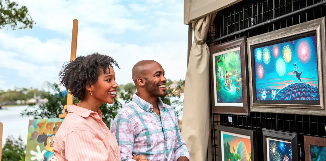 Two people happily viewing colorful framed artwork displayed outdoors at an art fair or exhibition