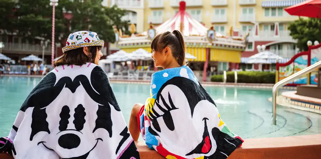 Two girls with colorful Disney character towels sitting by a pool at a resort with a carousel in the background