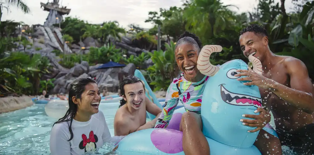 Group of diverse friends smiling and playing with a cartoonish blue inflatable dragon in a water park with lush greenery and rocky formations in the background