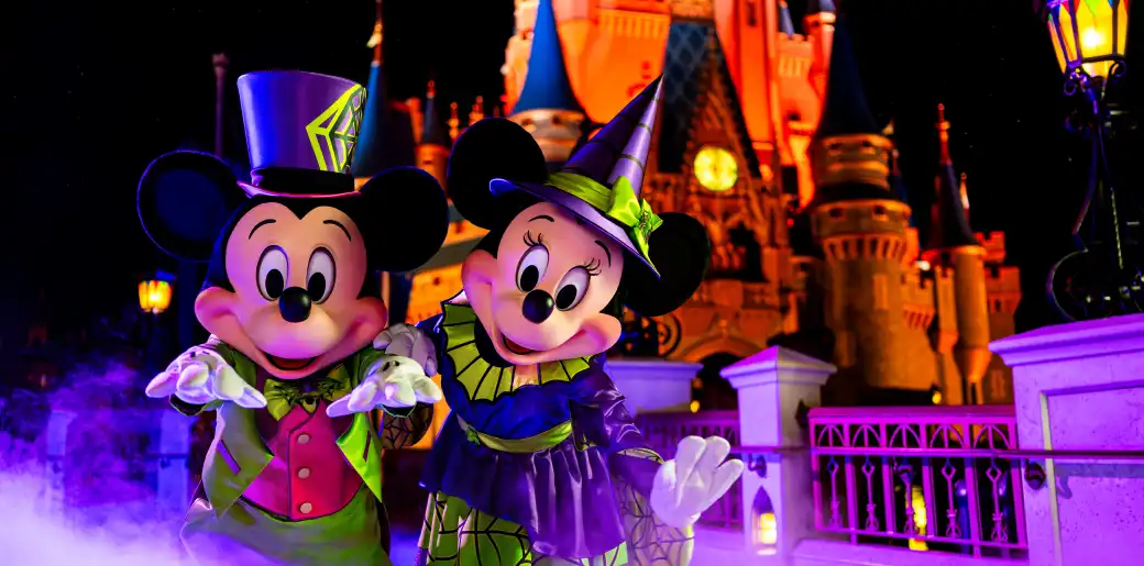 Mickey Mouse and Minnie Mouse dressed as witches at a nighttime Halloween event outside a Disney castle with purple and orange lighting.