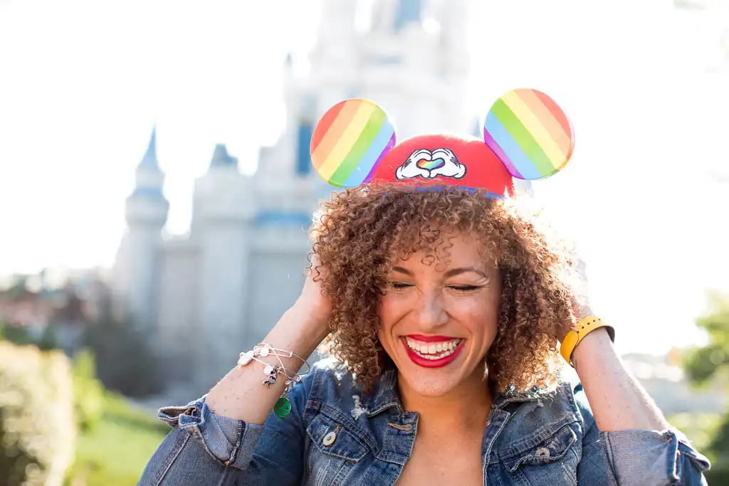 Woman smiling outdoors wearing rainbow Disney ear headbands and a red hat with a rainbow cloud design and a rainbow flag on the background castle.