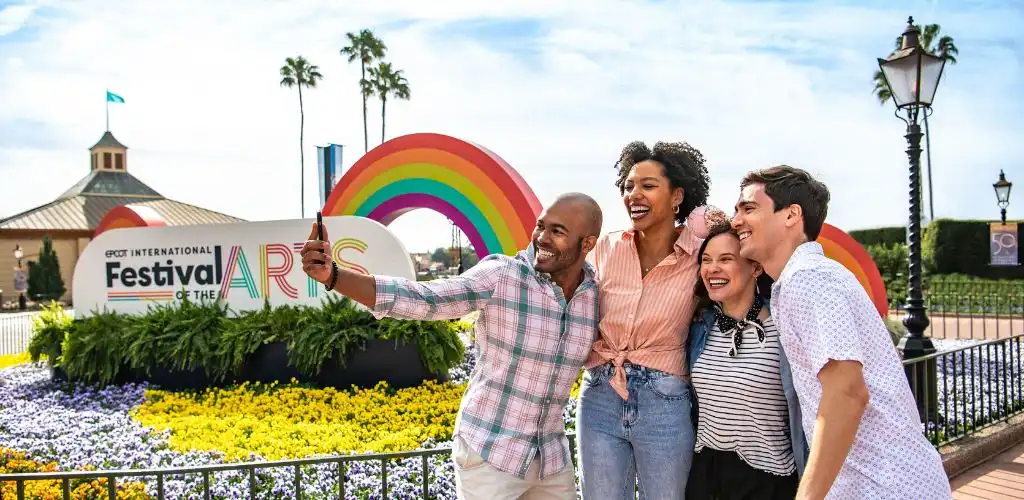 Group of diverse friends smiling and taking a selfie at an outdoor colorful festival with rainbow decoration and flowers.