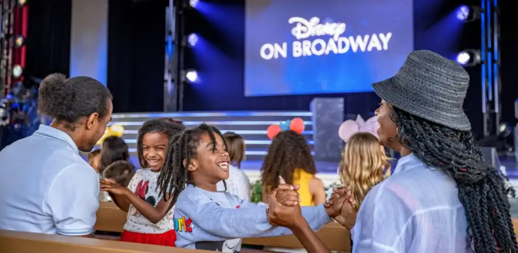 Group of children and adults enjoying a performance at Disney on Broadway stage with bright lights and colorful costumes