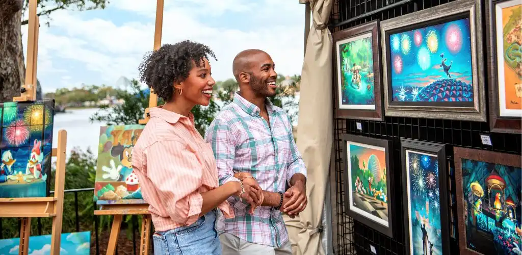 Two people smiling and enjoying colorful framed artwork at an outdoor art gallery or exhibition near a body of water.