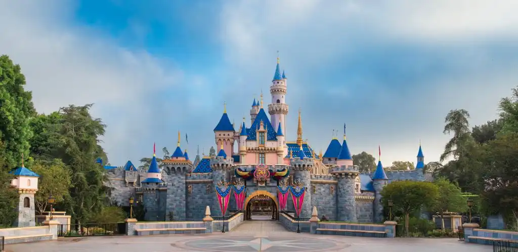 Disney castle with blue spires and pink accents surrounded by greenery and clear skies