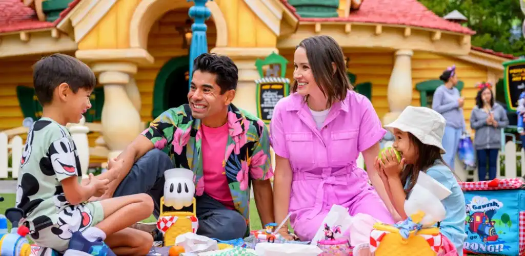 A family enjoying a picnic outdoors in front of a colorful playhouse, with children laughing and eating together on a bright, sunny day.