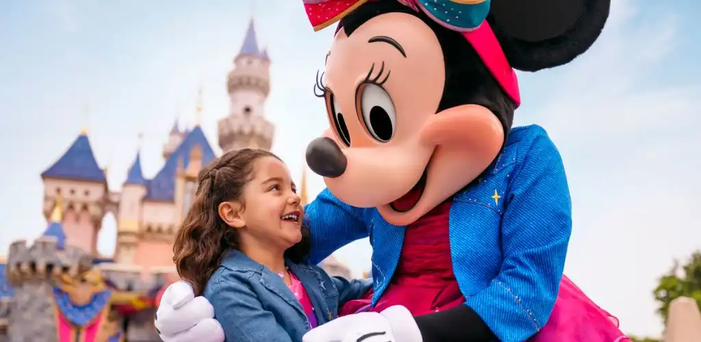 A girl smiling and hugging Minnie Mouse character in front of Sleeping Beauty Castle at Disneyland.