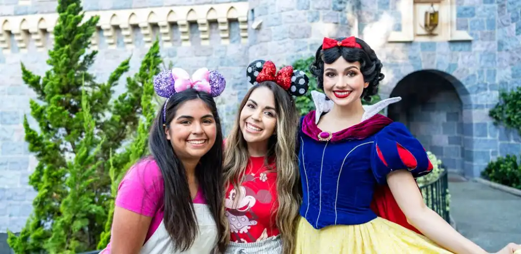 Three women dressed as Disney characters smiling outdoors with a stone building and greenery in the background. class=