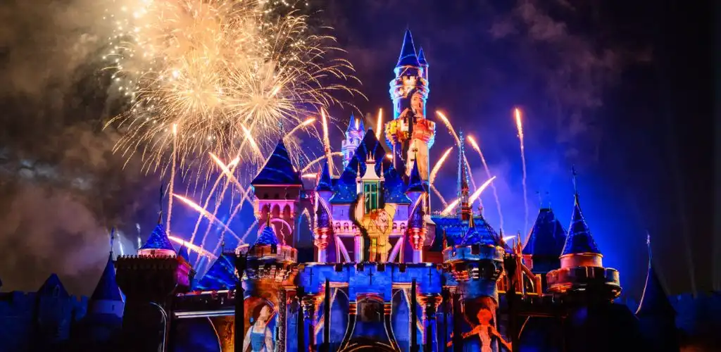 A brightly lit Disney castle at night with colorful fireworks exploding in the sky above.