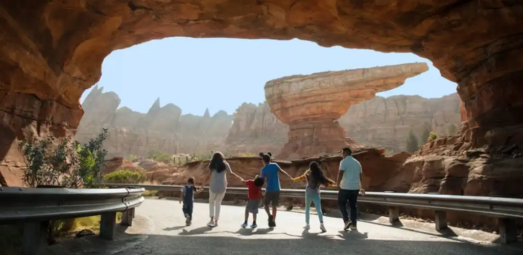 A family walks holding hands through a desert landscape with red rock formations and a blue sky behind them.