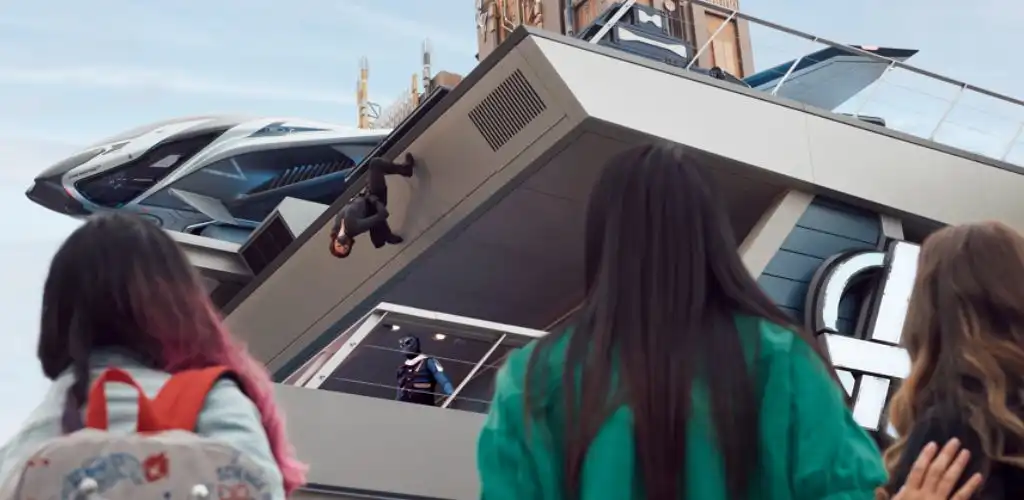 Three people stand outside a building looking up, with a man hanging upside down from the balcony above. Modern architecture and a sleek car are visible.