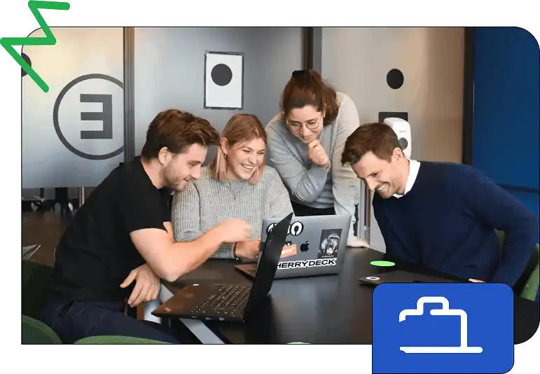 Four people gather around a table looking at a laptop and smiling during a collaborative activity inside a modern office space.