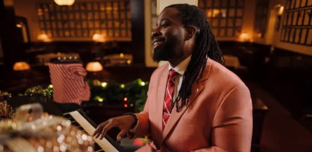 A man in a pink suit playing the piano in a warmly lit, festive environment with Christmas decorations and wrapped gifts.