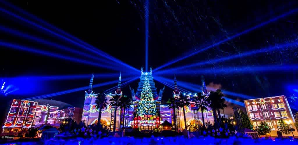 Nighttime holiday light display featuring a large decorated Christmas tree, colorful projections on buildings, palm trees, and laser beams in the sky