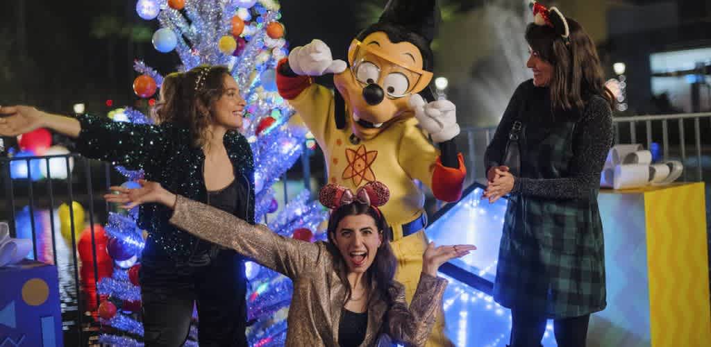 Three women and a person in a Mickey Mouse costume celebrate around a decorated Christmas tree at night with festive lights.