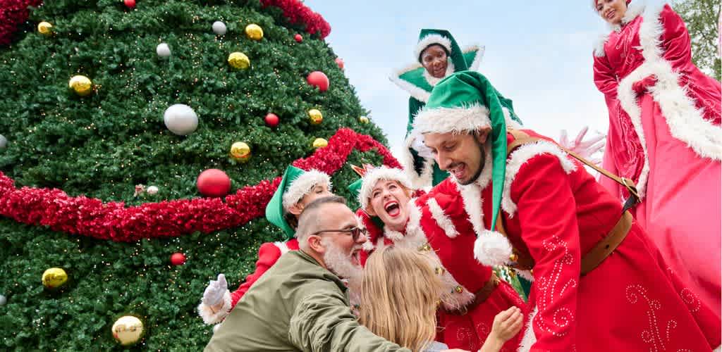 People gather outdoors around a decorated Christmas tree, smiling and laughing in festive costumes including elves, Santa, and holiday-themed clothing.
