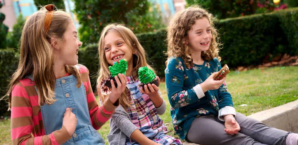 Three girls sitting outdoors sharing colorful cupcakes and smiling together.