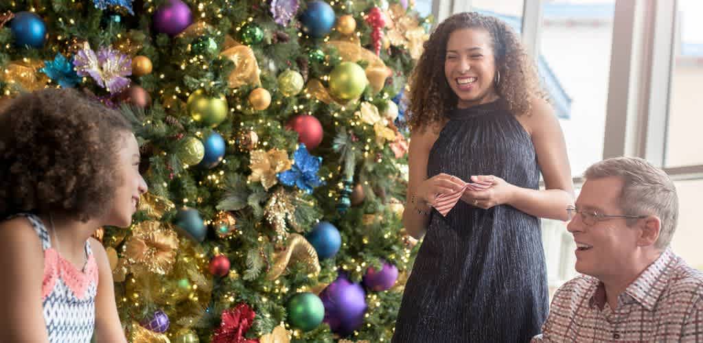 Three people smiling and talking near a decorated Christmas tree with colorful ornaments and lights.