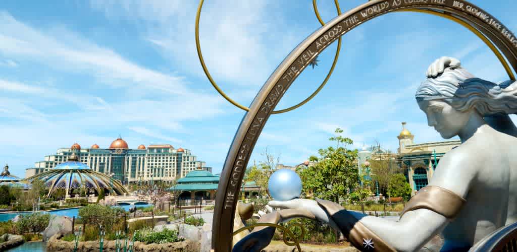 Statue of a woman with flowing hair holding a glowing orb, with a large themed hotel in the background and blue sky overhead.