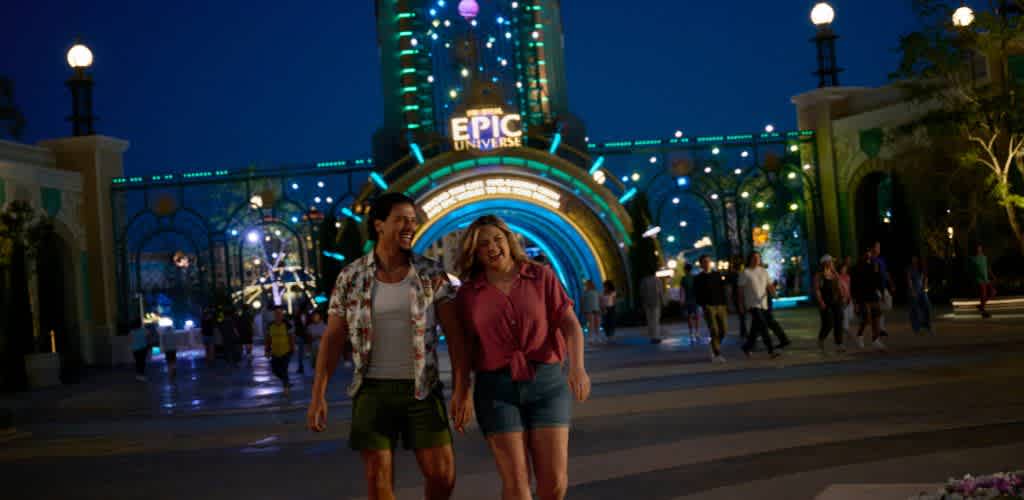 Two women smiling and walking together at night in front of an illuminated amusement park entrance with colorful lights and crowd of visitors.