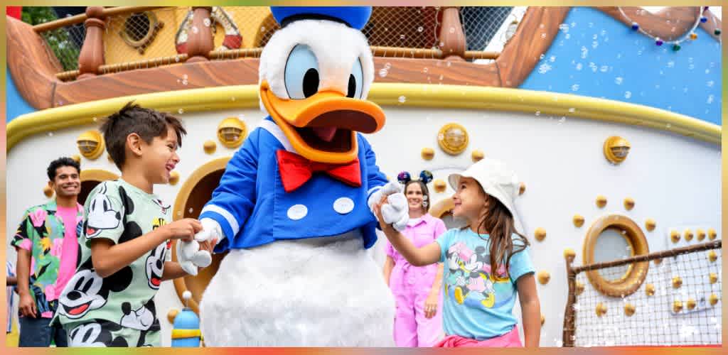 Children with play uniforms and a girl with a hat interact happily with a person dressed as Donald Duck in front of a large ship structure at a theme park.