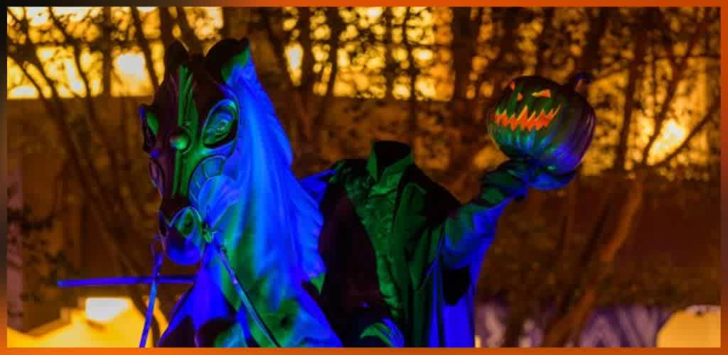 Person in a green costume wearing a glowing mask holding a carved pumpkin with a face in a nighttime outdoor setting with trees and warm lighting.