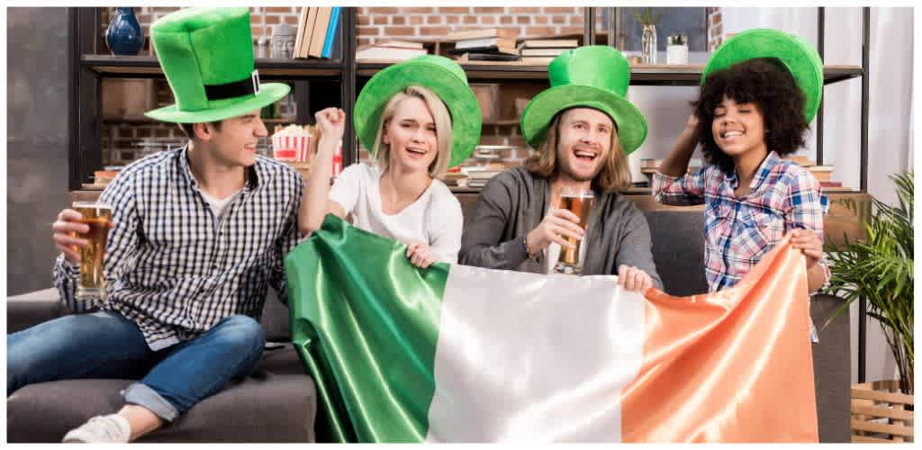 Group of four friends wearing green leprechaun hats celebrating with drinks and holding an Irish flag indoors