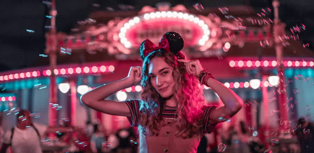 A young woman with curly hair wearing Minnie Mouse ears at a brightly lit amusement park at night with pink and blue neon lights.