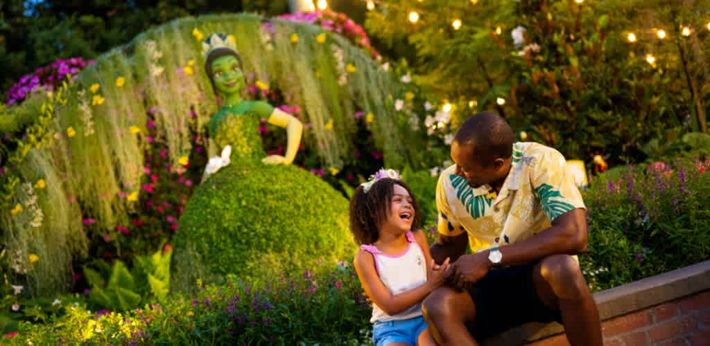 A father and daughter smiling and laughing together in a colorful garden with a large topiary sculpture of a woman in a dress.