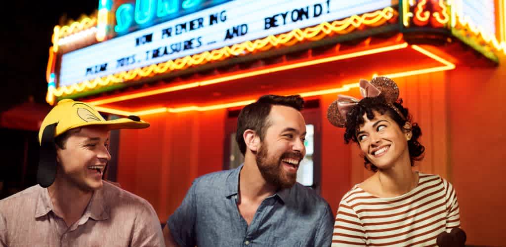 Three friends smiling and having fun at a carnival or amusement park night, wearing playful hats including a yellow cartoon character hat and animal ears.